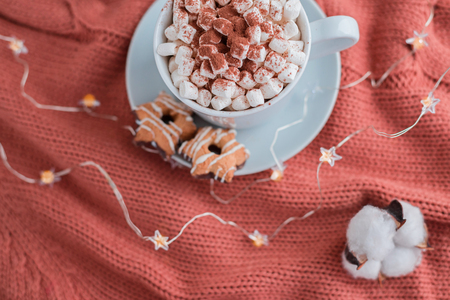 Coffee cup with marshmallow and stars cookies on warm knitted coral blanket, christmas lights and cotton flowers. Cozy and moody concept.の写真素材