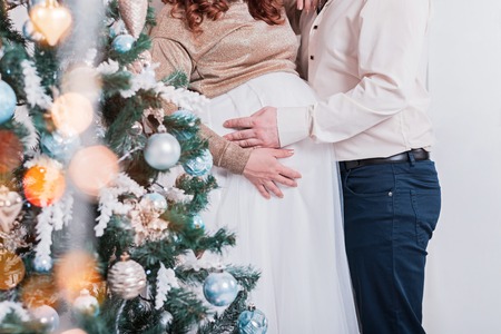 Close up of parents hands touching pregnant belly near the christmas tree. Cropped.の写真素材