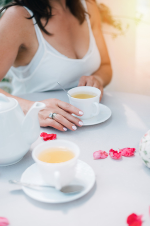 Tea cups and womans hands holding one cup on table with rose petals, top view.の写真素材