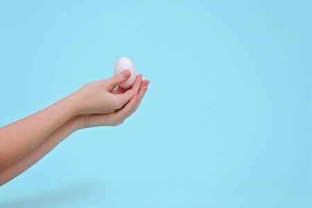 Woman's hands with white egg on pastel blue background. Easter concept. Copy space.の写真素材
