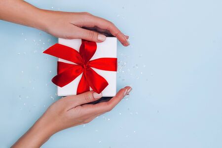 Woman's hands with manicure hold gift box with red ribbon on pale blue background with glitters. Festive concept. Place for text.の写真素材