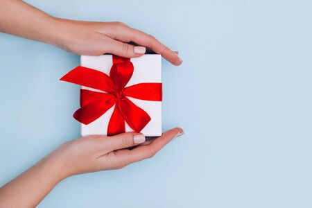 Womans hands with manicure hold gift box with red ribbon on pale blue background. Festive concept.の写真素材