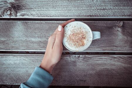 Woman's hand in blue sweater holding cup of cappuccino on the grey wooden table. Top view.の写真素材