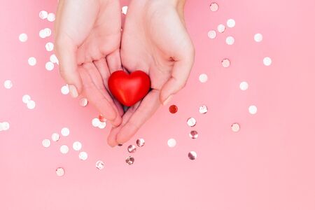Womans hands with manicure hold red heart on pale pink background with glitters. Festive concept.の写真素材