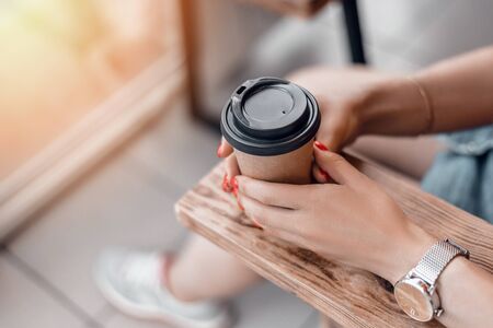 Paper coffee cup to go in womans hands with red manicure while sitting in cafe.の写真素材