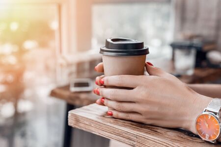 Paper coffee cup to go in womans hands with red manicure while sitting in cafe.の写真素材