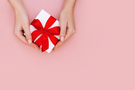 Woman's hands with manicure hold gift box with red ribbon on pale pink background. Festive concept. Place for text.の写真素材