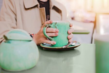 Big mint green tea cup in woman's hands with pastel manicure while sitting in cafe. Cropped.