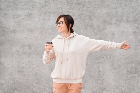 Young pretty woman in white hoody and glasses with paper coffee cup to go in her hands, happy and smiling. Mockup.の写真素材