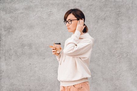 Young pretty woman in white hoody and glasses with paper coffee cup to go in her hands. Mockup.の写真素材