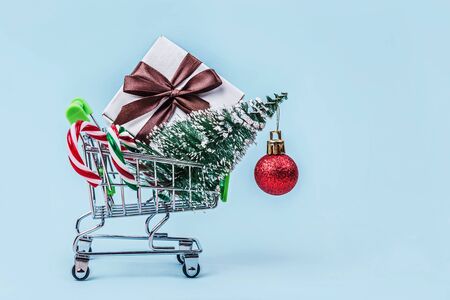 Christmas tree, gift box and candy canes in miniature shopping cart with on pastel blue background with copy space. Christmas shopping concept.の写真素材
