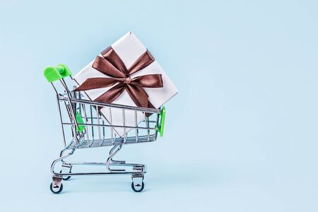 Gift box with brown bow in miniature shopping cart on pastel blue background with copy space. Shopping concept.の写真素材
