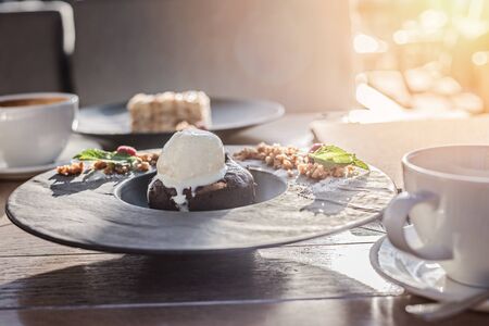 Cake with ice-cream in black plate and white big cup of delicious coffee on wooden table in sunset light. Lifestyle view.の写真素材