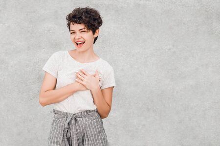 Charming young woman with creative hairstyle is smiling and posing with joyful face hold her hands near heart.の写真素材