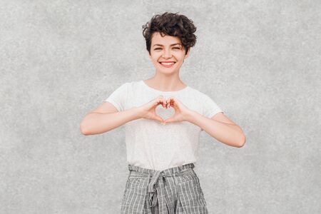 Charming young dark-haired woman with creative hairstyle is smiling and with joyful face makes heart with fingers.の写真素材