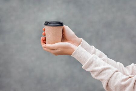 Woman in white hoody holds paper coffee cup to go in her hands on grey background, cropped. Mockup. Copy space.の写真素材