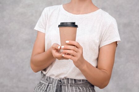 Woman in white t-shirt holds paper coffee cup to go in her hands on grey background, cropped. Mockup. Copy space.の写真素材