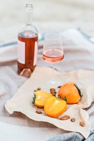 Picnic on the beach with persimmons, almond and bottle of rose wine on beige blanket. Lifestyle view.の写真素材