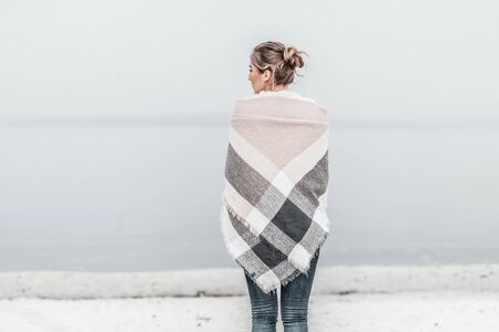 Pretty blonde woman wrapped in warm blanket standing on the cold autumn beach. Copy space.の写真素材
