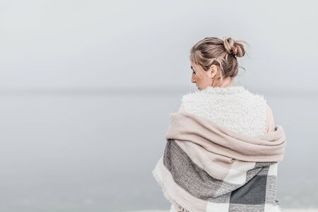 Pretty blonde woman wrapped in warm blanket standing on the cold autumn beach. Copy space.の写真素材