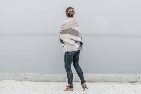Pretty blonde woman wrapped in warm blanket standing on the cold autumn beach.の写真素材