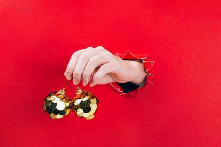 Female hand holding two golden Christmas tree baubles through a hole in red background. Minimal Christmas, New year concept.の写真素材