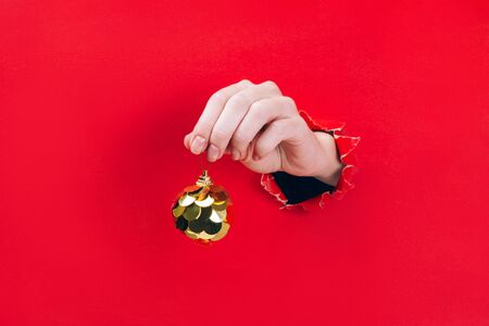 Female hand holding golden Christmas tree bauble through a hole in red background. Minimal Christmas, New year concept.の写真素材