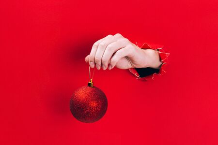 Female hand holding glittering red Christmas tree bauble through a hole in red background. Minimal Christmas, New year concept.の写真素材