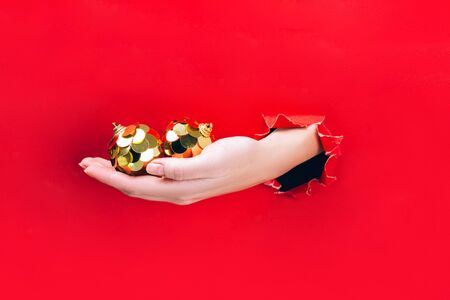 Female hand holding two golden Christmas tree baubles through a hole in red background. Minimal Christmas, New year concept.の写真素材