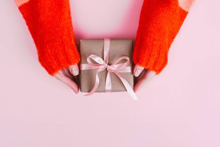 Woman's hands in warm knitted sweater with manicure hold gift box wrapped with craft paper and pink ribbon on pale pink background. Festive concept. Place for text.の写真素材