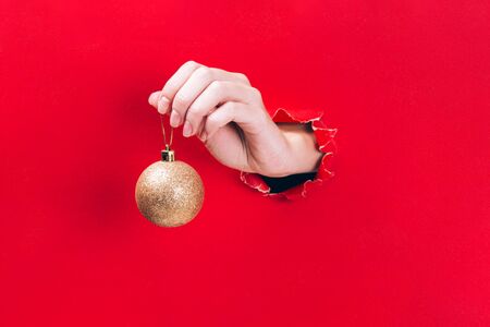 Female hand holding golden Christmas tree bauble through a hole in red background. Minimal Christmas, New year concept.の写真素材