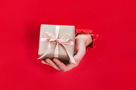 Male hand holding gift box wrapped in craft paper with pink ribbon through a hole in red background. Minimal concept.の写真素材