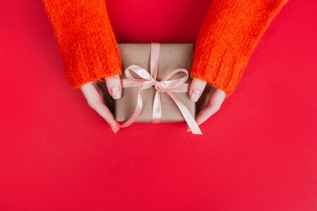 Woman's hands in warm knitted sweater with manicure hold gift box wrapped with craft paper and pink ribbon on red background. Festive concept. Place for text.の写真素材