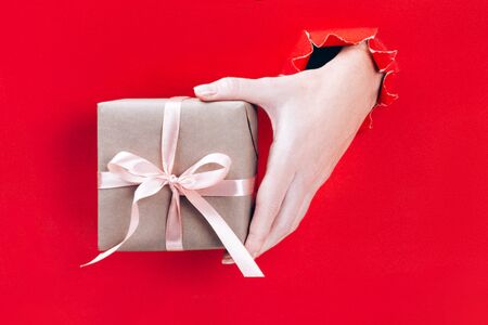 Female hand holding gift box wrapped in craft paper with pink ribbon through a hole in red background. Minimal festive concept.の写真素材