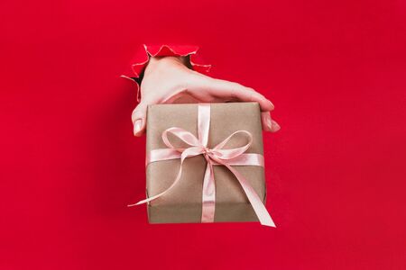 Female hand holding gift box wrapped in craft paper with pink ribbon through a hole in red background. Minimal festive concept.の写真素材