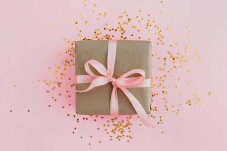 Gift box wrapped in craft paper with pink ribbon on pale pink background with glitters. Festive concept, top view.の写真素材
