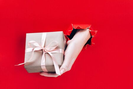 Female hand holding gift box wrapped in craft paper with pink ribbon through a hole in red background. Minimal festive concept.の写真素材