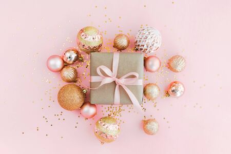 Gift box wrapped in craft paper with pink ribbon on pale pink background with glitters and golden Christmas tree balls. Festive concept, top view.の写真素材