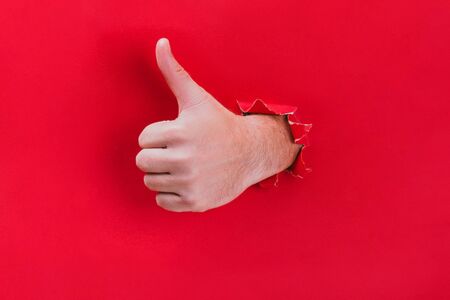 Male hand breaks through the red paper background and shows his thumb up. Minimal concept.の写真素材