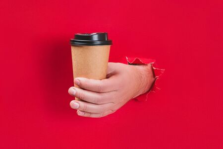 Male hand holding craft paper coffee cup through a hole in red background.の写真素材