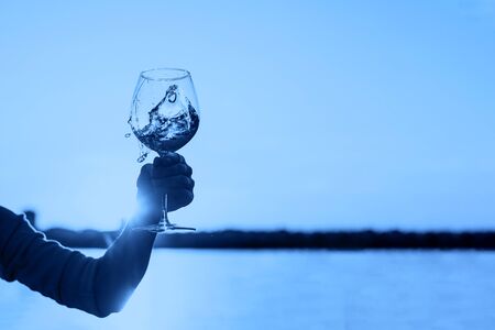 Glass of wine in woman's hand against the sunset sky toned blue color. Cropped.の写真素材