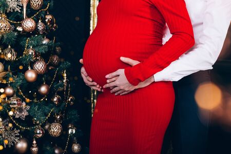 Parents hands touching pregnant belly near christmas tree decorated with golden toys. Cropped, close up.の写真素材