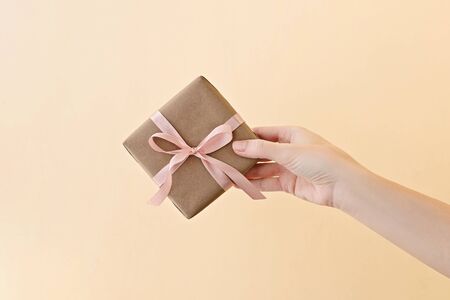 Female hand holding gift box wrapped in craft paper with pink ribbon through on pale beige background.の写真素材