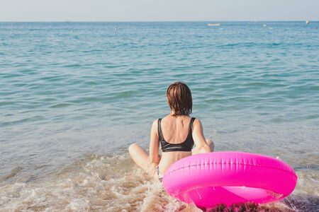 Young woman with pink swimming ring is sitting on the at beach sea in sunny day. Summer vacation concept. Back view.の写真素材