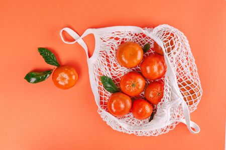 Fresh ripe mandarins with green leaves in white cotton mesh bag on orange background. Flat lay, top view, copy space.の写真素材