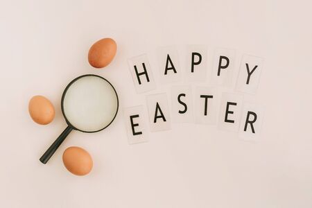 Magnifying glass among brown eggs and words Happy easter on beige background. Easter concept.