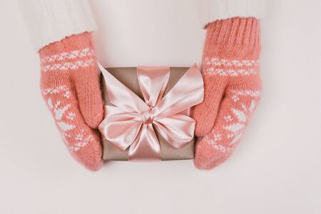 Woman's hands in warm knitted mittens hold gift box wrapped with craft paper and pink ribbon on white background. Festive concept. Place for text.の写真素材