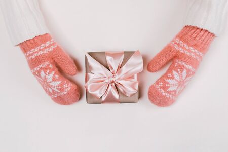 Woman's hands in warm knitted mittens hold gift box wrapped with craft paper and pink ribbon on white background. Festive concept. Place for text.の写真素材