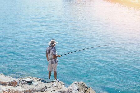 Fisherman in big hat is fishing on sea rocks alone in sunny day. Back view.の写真素材