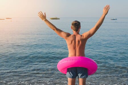Happy man in pink with a big inflatable ring on the sea beach in summer sunny day. Back view.の写真素材